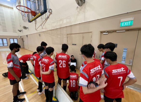 A group of youth in red SST jerseys huddle with a coach in an indoor basketball gym near an exit door.